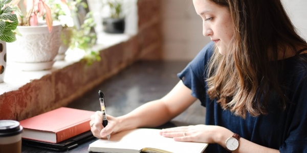 woman-writing-in-journal-as-she-sits-near-window-with-plants-on-ledge